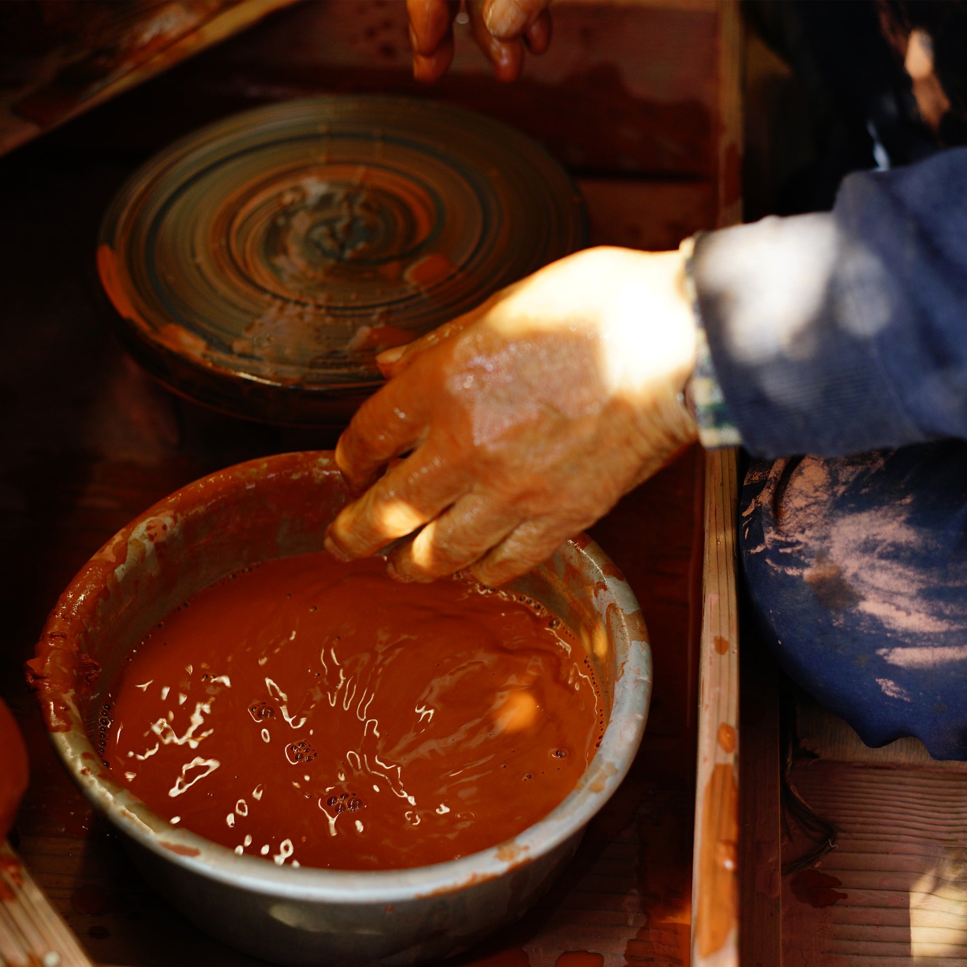 Artist working with clay on a pottery wheel, with a bowl of reddish-brown liquid nearby.