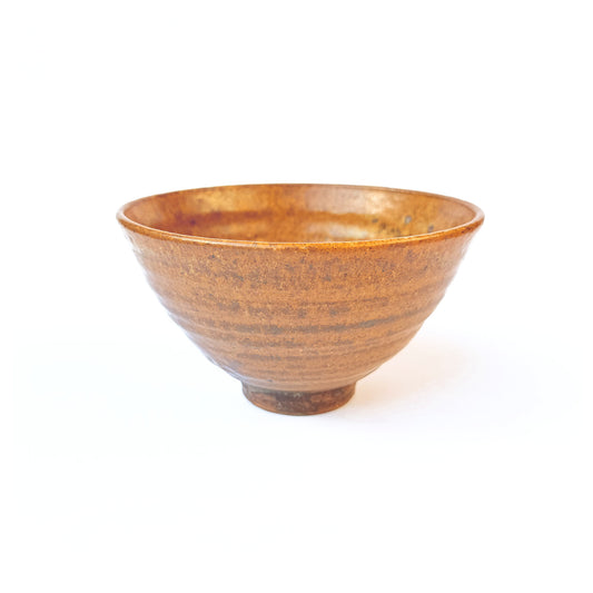 handcrafted RAKU ceramic matcha bowl with a warm, burnished glaze showing gradients of amber, bronze, and charcoal, and gentle spiral ridges inside.
