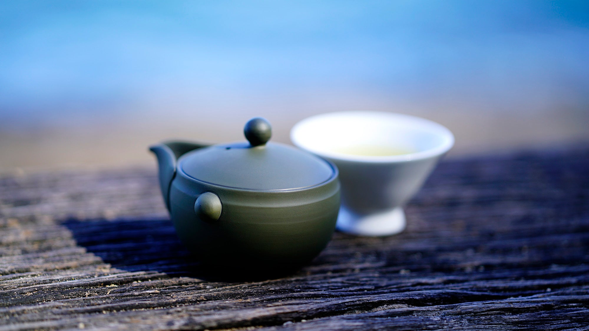 Green handleless teapot and white cup on a wooden surface with a blurred natural background