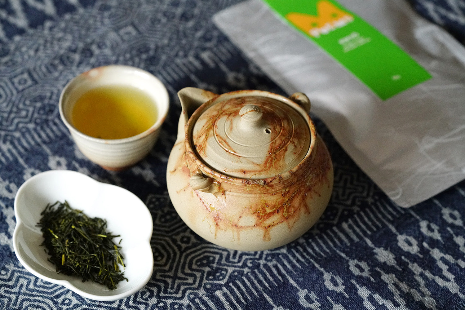 Tea set with a teapot, cup, and bowl of tea leaves on a patterned fabric background.