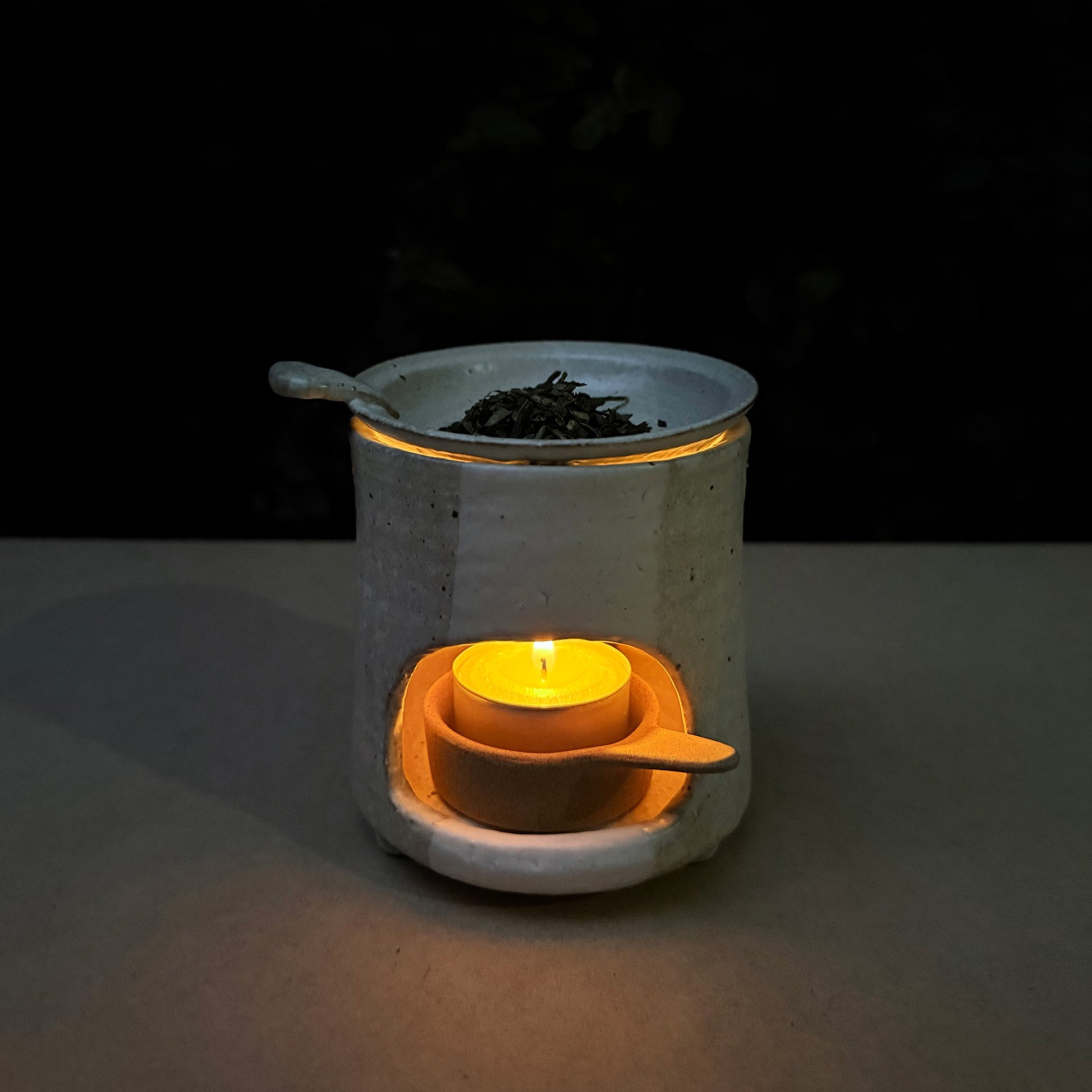 Ceramic tea warmer with a lit candle inside, surrounded by loose tea leaves on a dark background.