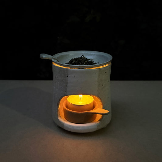 Ceramic tea warmer with a lit candle inside, surrounded by loose tea leaves on a dark background.